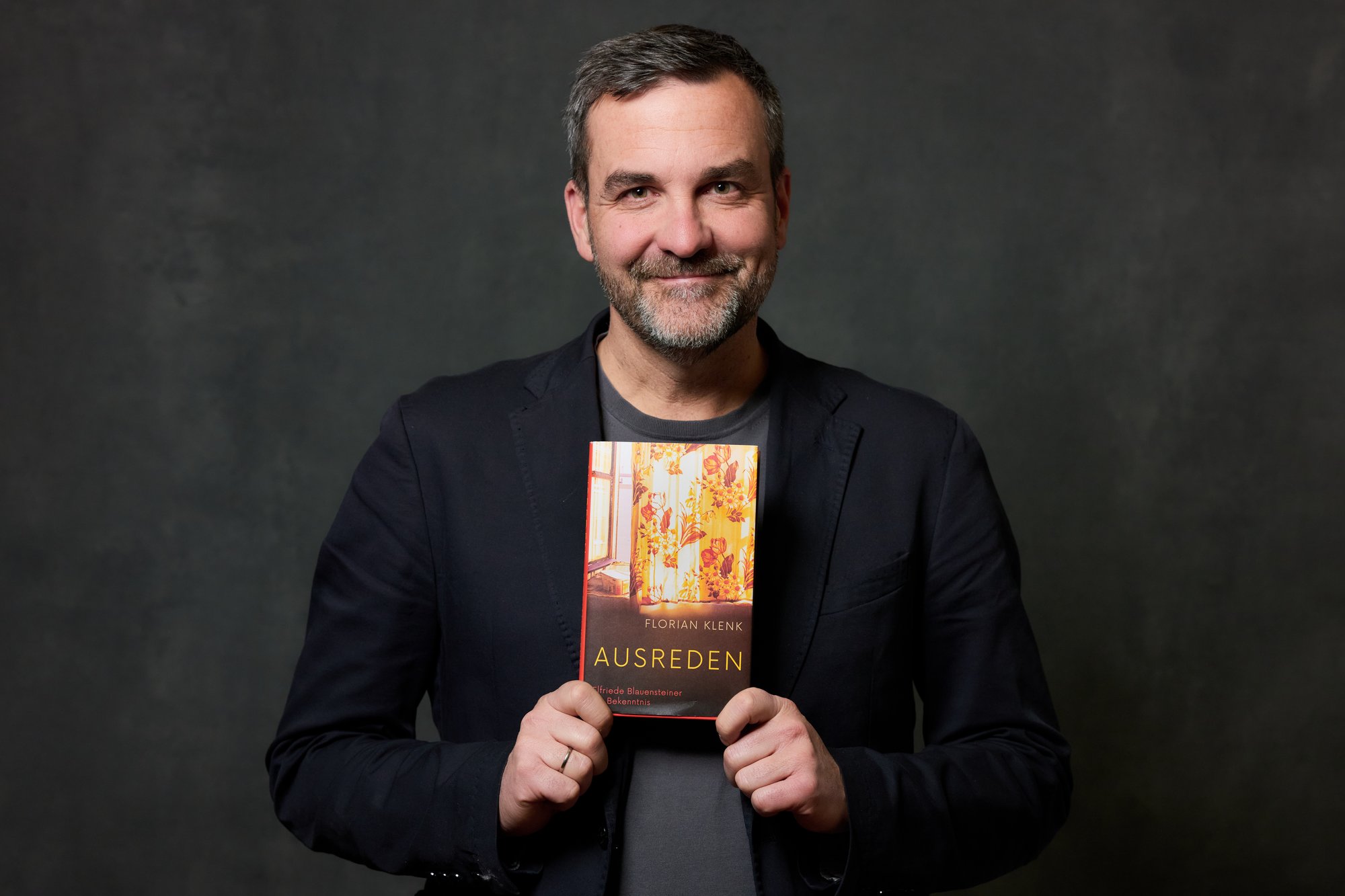 Portrait von Florian Klenk, der lächelnd sein Buch "Ausreden. Elfriede Blauensteiner - Ein Bekenntnis" vor der Brust hält.